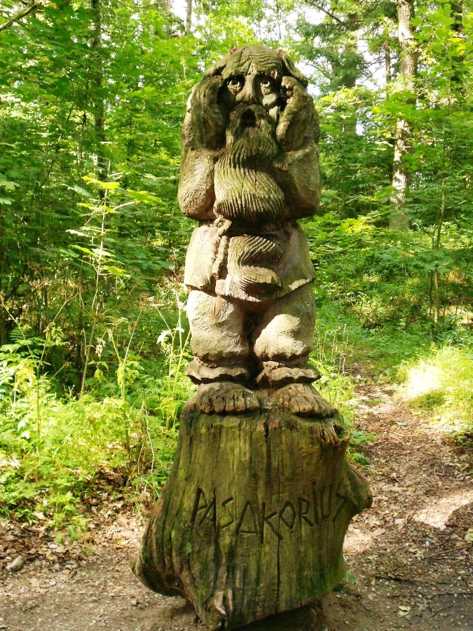 Eine Holzskulptur eines mystischen Wesens mit einem Bart, das die Hände an den Kopf legt, umgeben von Bäumen und Pflanzen in einem Wald.
