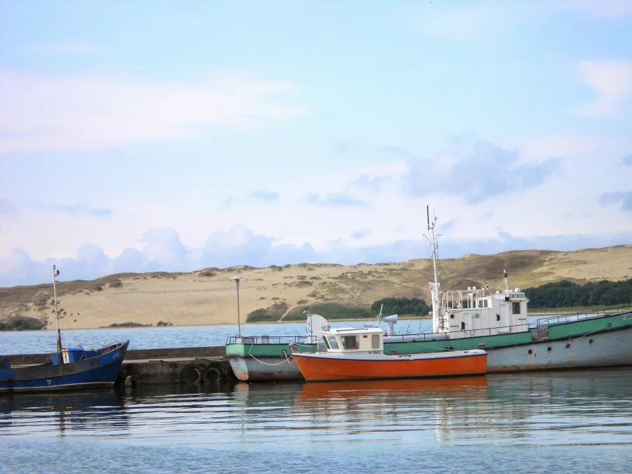 Boats docked by a calm body of water with sandy dunes in the background and a blue sky.