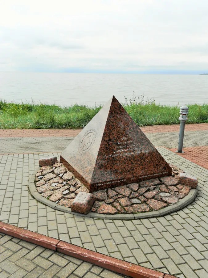 Ein pyramidenförmiges Denkmal aus Stein steht auf einem gepflasterten Weg am Ufer, umgeben von Gras und dem Wasser im Hintergrund.