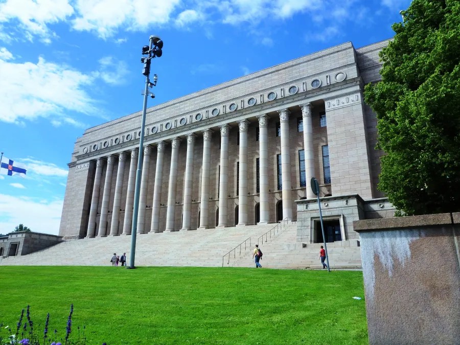 A view of the Finnish Parliament House in Helsinki, showcasing its grand architecture with large columns and steps, surrounded by green grass and a blue sky.