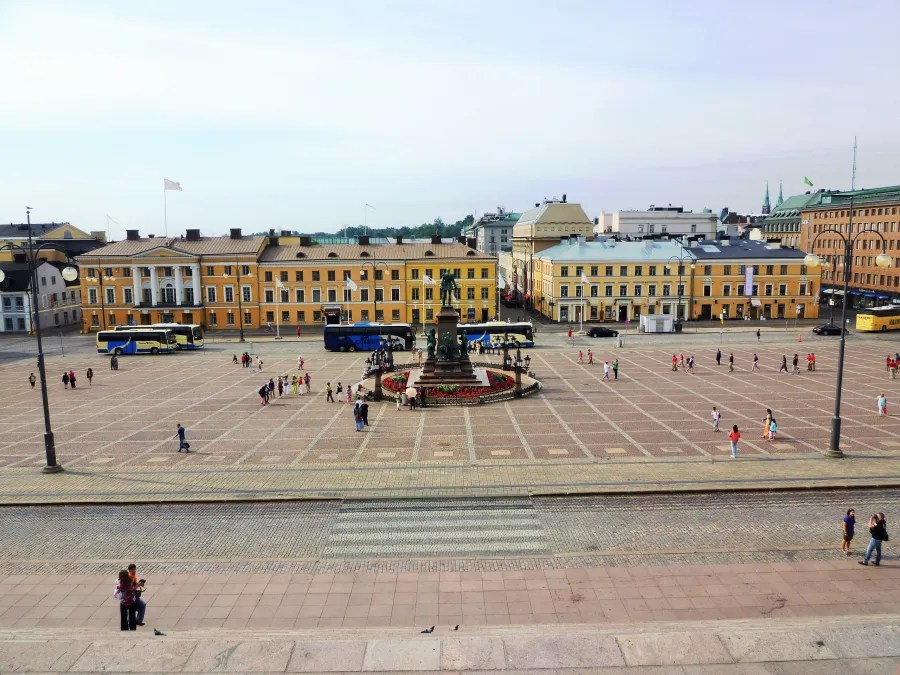A view of a large square in Helsinki with a central monument surrounded by yellow buildings and people walking around. Buses are parked at the edge of the square.
