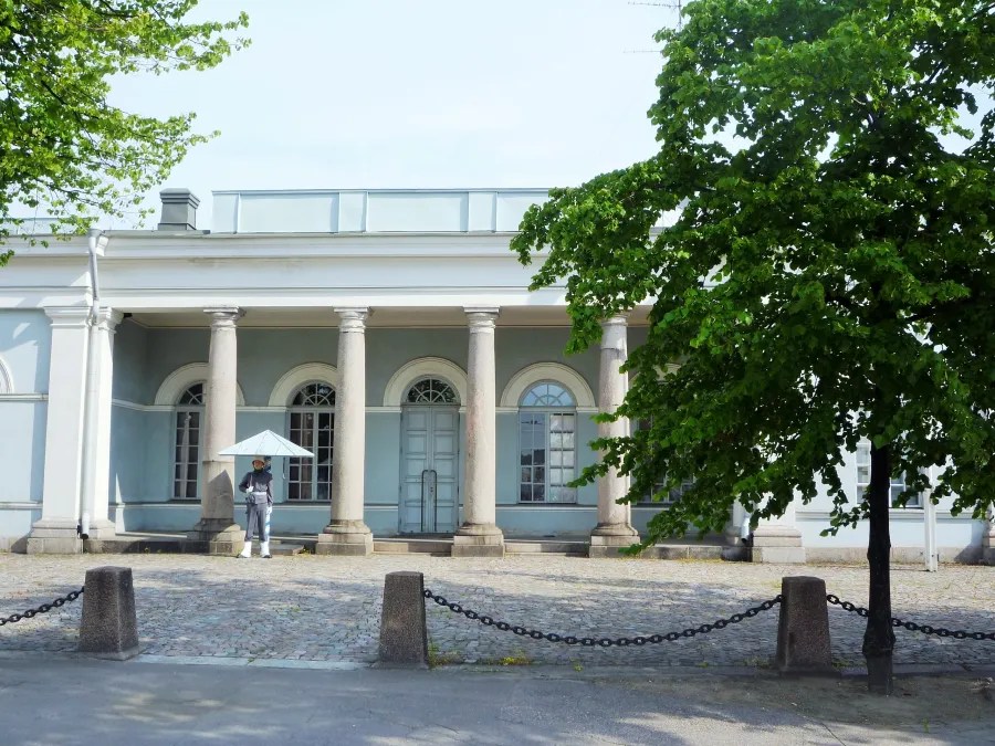 Ein Gebäudefront mit Säulen und einem Mann, der einen Sonnenschirm hält, umgeben von Bäumen und einem gepflasterten Platz.