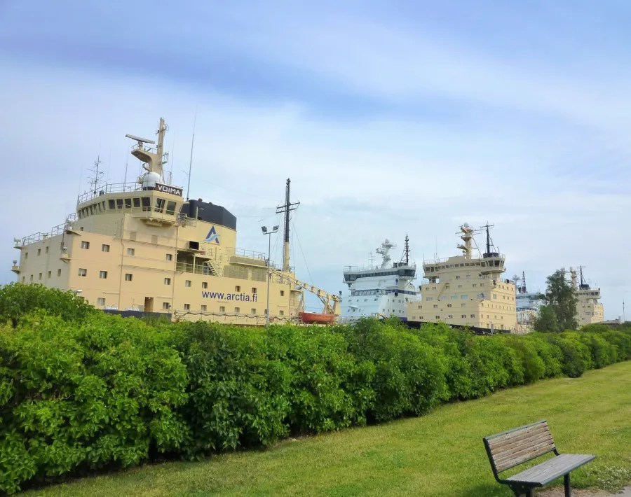 Three icebreaker ships docked beside a green hedge, with a bench in the foreground and a cloudy sky above.