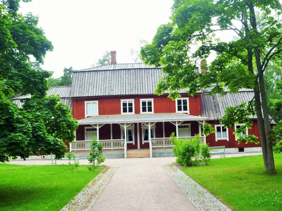 A red wooden house with a pitched roof, surrounded by trees and greenery, featuring a porch and large windows.