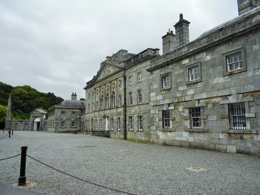 Ansicht des Powerscourt House mit grauem Steinbau und Gartenanlagen im Vordergrund, umgeben von Bäumen unter einem bewölkten Himmel.
