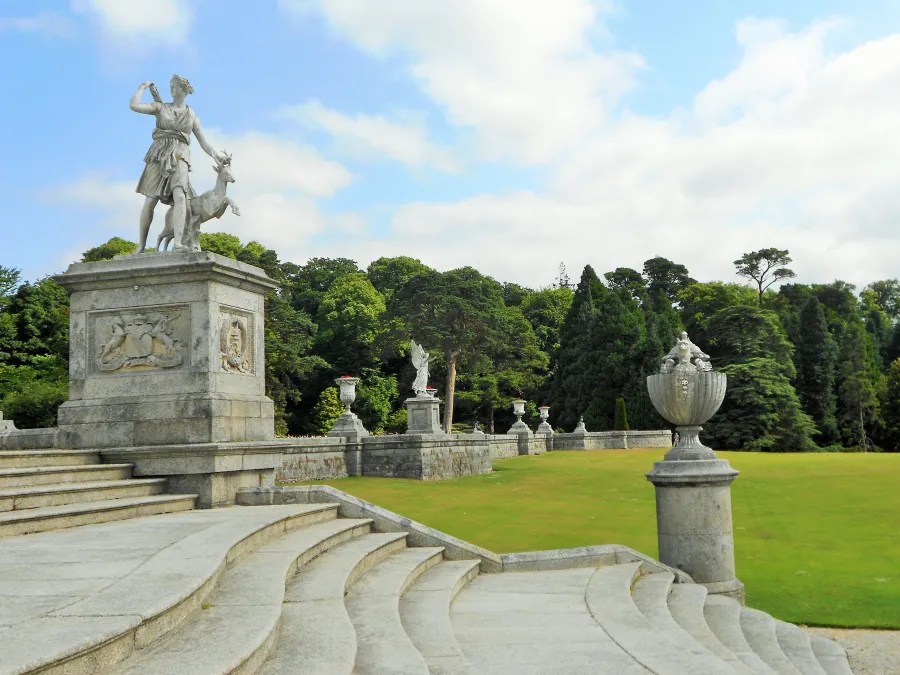 Blick auf die Treppe und die Statue eines Mannes mit einem Hund im Powerscourt House & Gardens, umgeben von üppigem Grün.