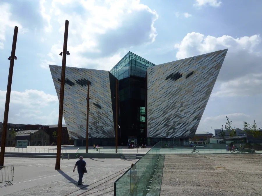 Das Titanic Belfast Gebäude mit seiner modernen Architektur und dem spitz zulaufenden Dach, umgeben von einem offenen Platz und einer klaren blauen Himmel.