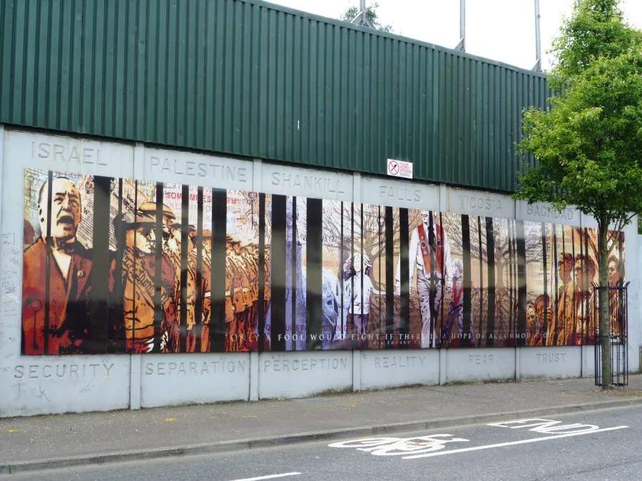 Wandgemälde an einer Mauer in Belfast, das verschiedene historische und politische Themen darstellt, mit menschenähnlichen Figuren und Texten zu Sicherheit, Trennung und Realität.
