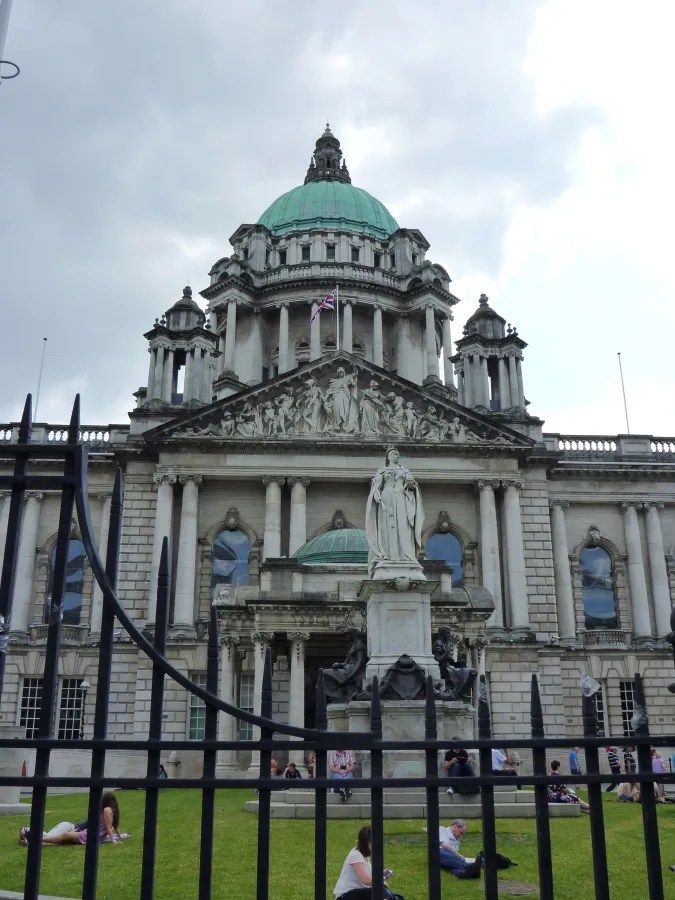Ansicht des Rathauses von Belfast mit einer Statue im Vordergrund und Menschen auf dem Rasen, bewölkten Himmel und grünem Dach.
