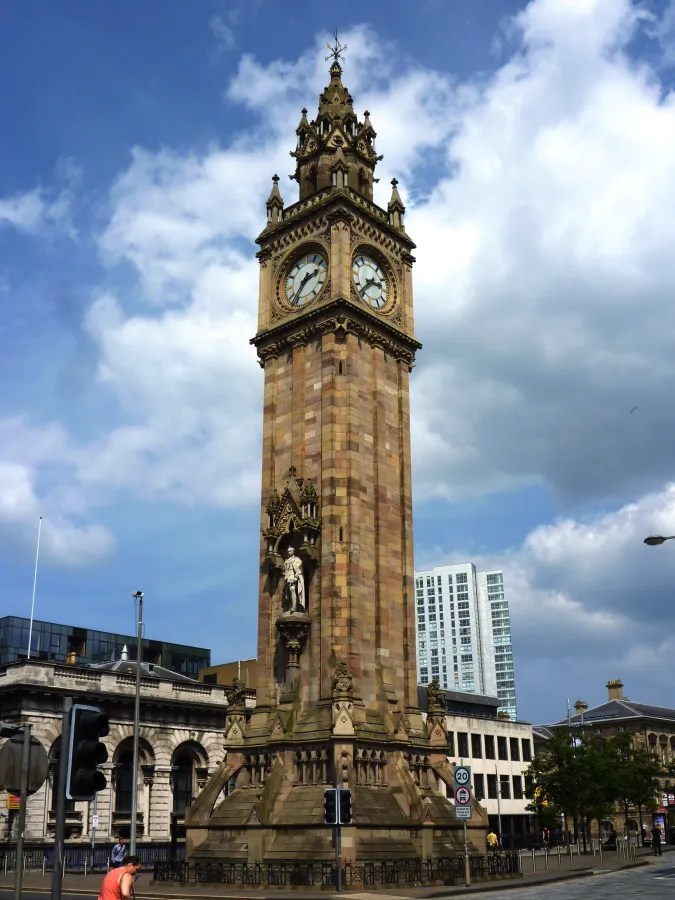 Ein majestätischer Uhrturm in Belfast mit detaillierter Architektur, umgeben von modernen Gebäuden und einem blauen Himmel mit Wolken.