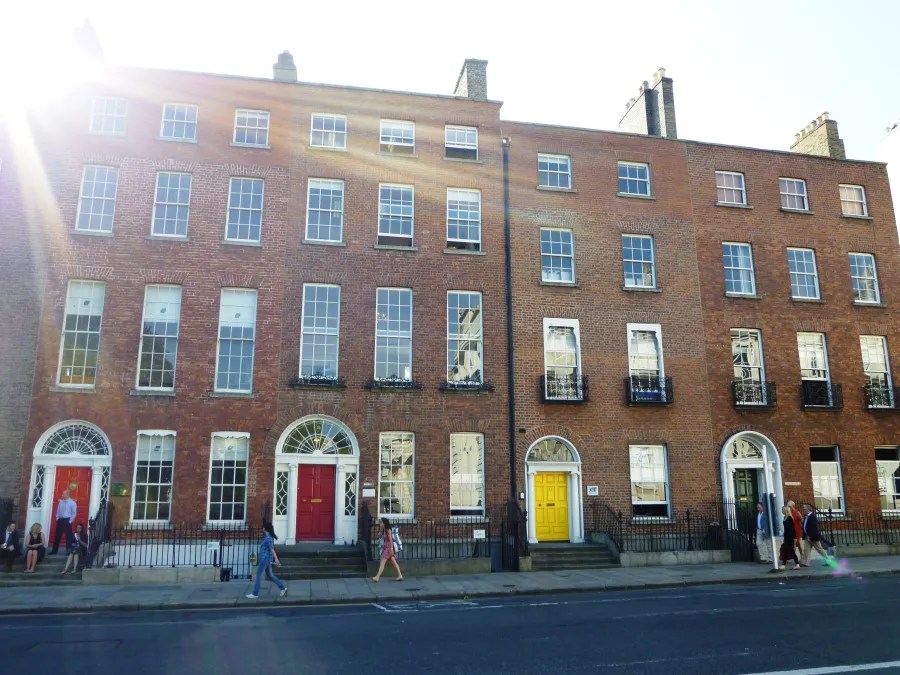 Eine Reihe von historischen roten Backsteinbauten mit mehreren Fenstern und bunten Türen in Dublin, Irland. Menschen gehen an den Gebäuden vorbei, während die Sonne scheint.