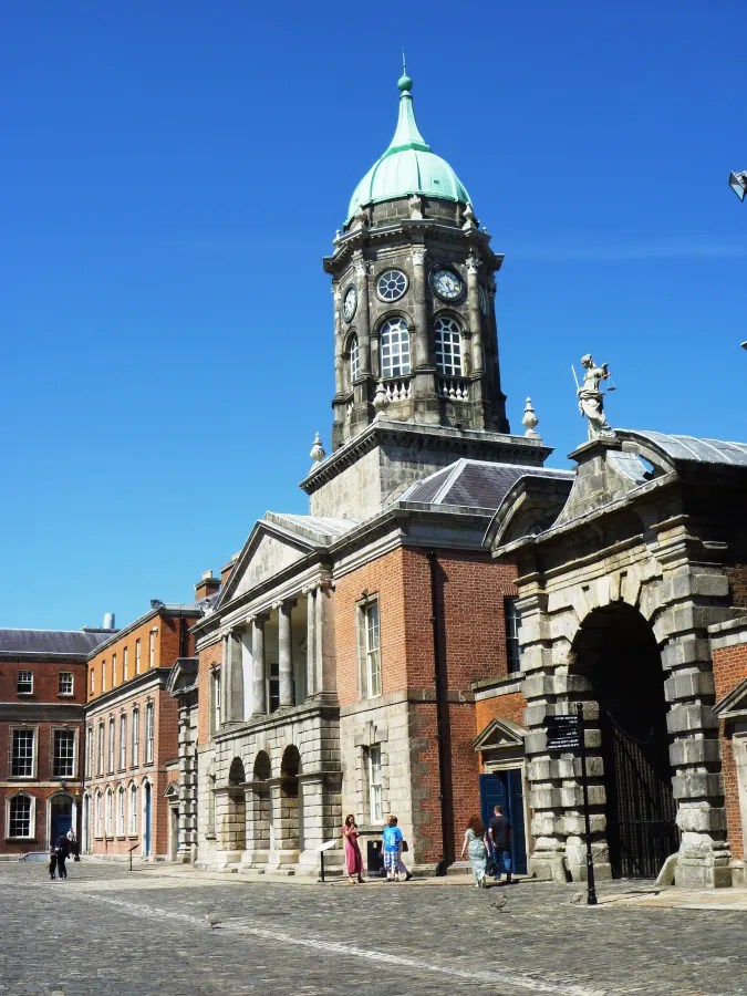 Blick auf ein historisches Gebäude mit einer markanten Turmhaube und Säulenfassade unter strahlend blauem Himmel, umgeben von einer gepflasterten Fläche und Besuchern.