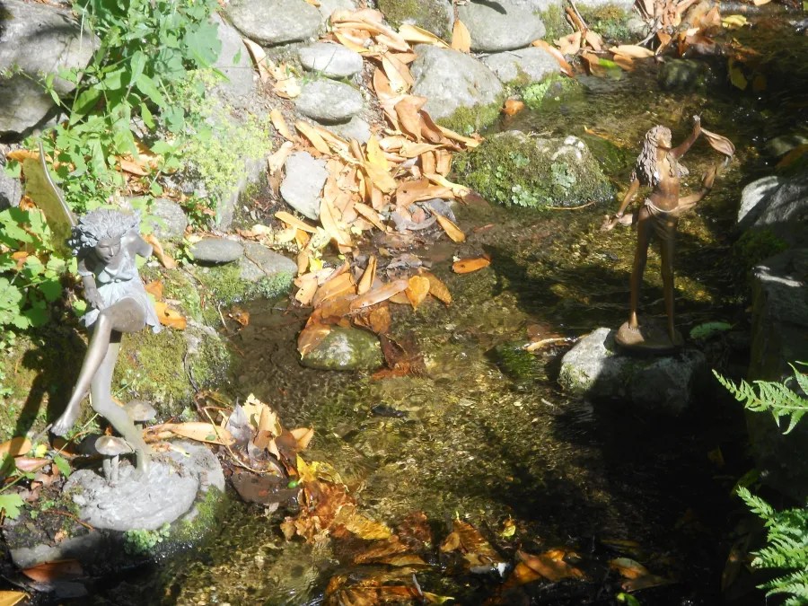 Eine Szene mit zwei stilisierten Figuren an einem kleinen Wasserlauf, umgeben von herbstlichen Blättern und Steinen.