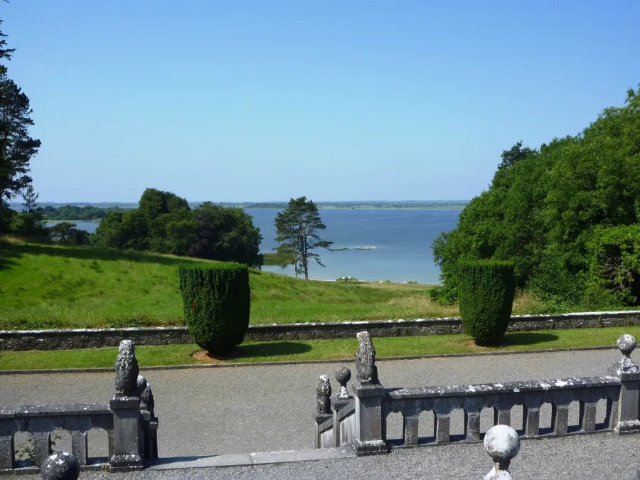 Blick auf einen ruhigen See umgeben von grünen Wiesen und Bäumen, mit einem Geländer im Vordergrund.
