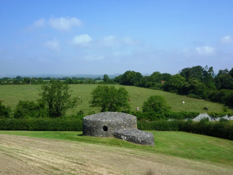 Blick auf eine ländliche Landschaft mit einer Steinhütte im Vordergrund, umgeben von grünen Wiesen und Bäumen unter einem klaren blauen Himmel.