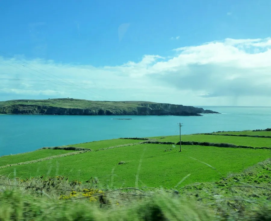 Blick auf die grüne Landschaft Irlands mit sanften Hügeln und einem ruhigen blauen Meer im Hintergrund, unter einem klaren blauen Himmel.