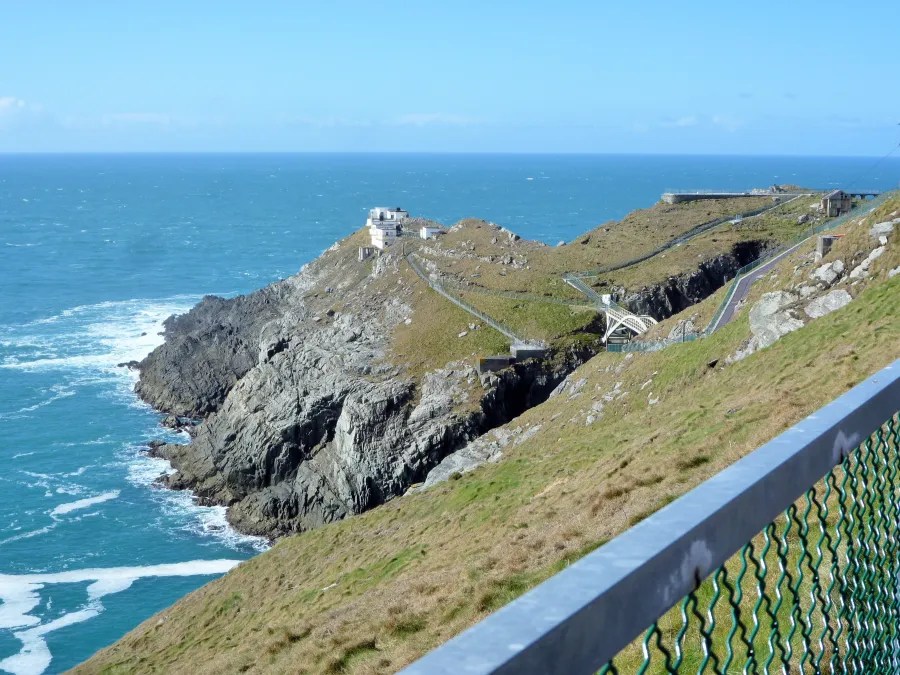 A scenic view of Mizen Head in Ireland, featuring rocky cliffs, lush green grass, and a lighthouse in the distance, alongside the blue ocean waves crashing against the shore.