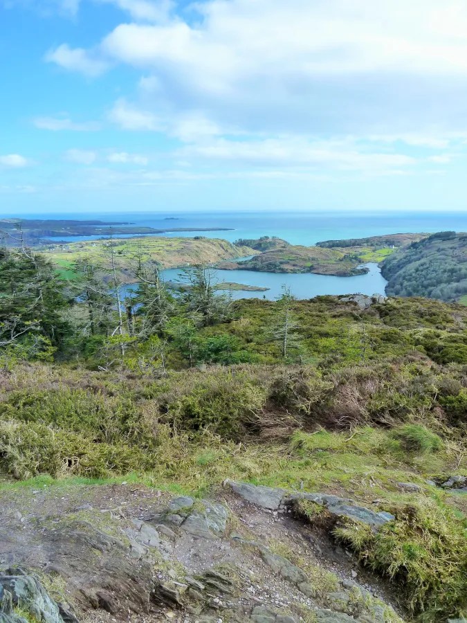 Blick auf eine grüne Landschaft mit einem ruhigen See und dem Atlantik im Hintergrund unter einem teilweise bewölkten Himmel.