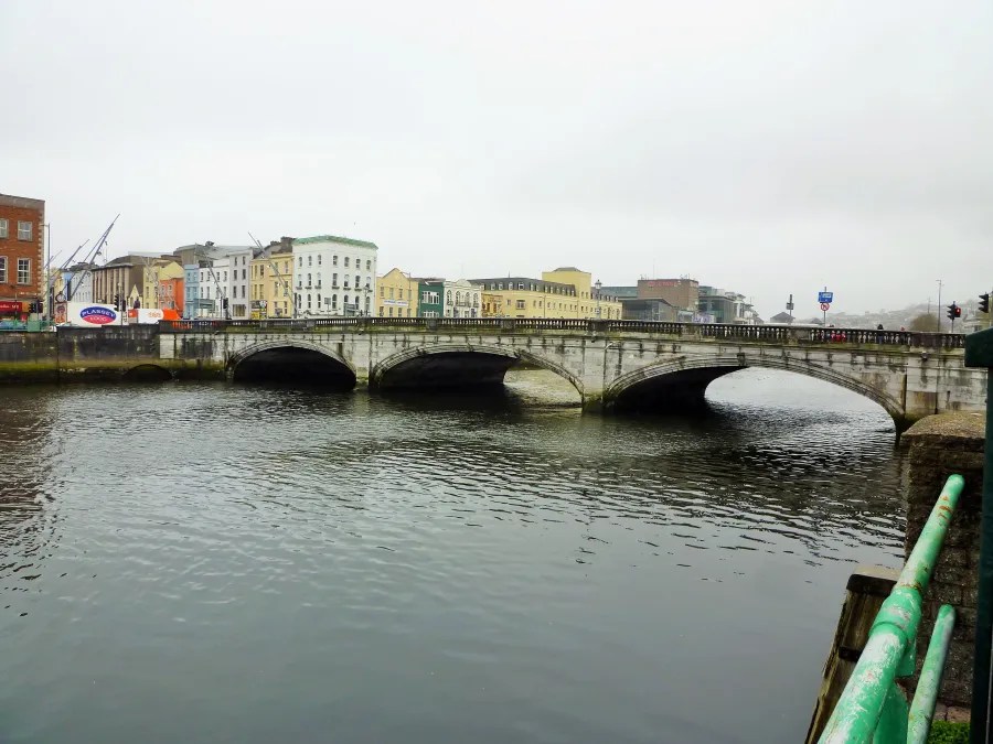 Ein Blick auf eine Brücke in Cork, Irland, mit bunten Gebäuden im Hintergrund und Wasser im Vordergrund, an einem bewölkten Tag.
