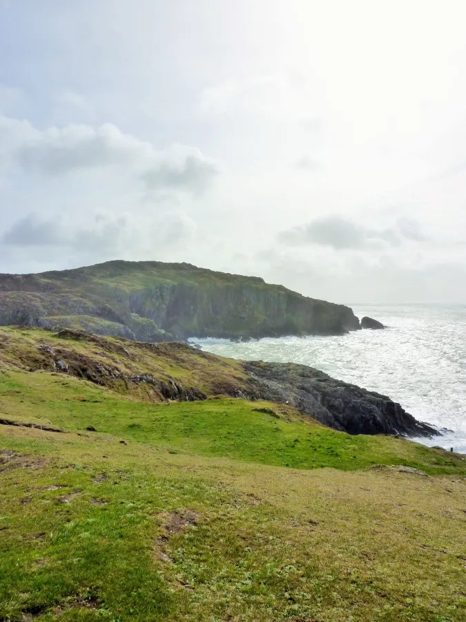 Blick auf eine grüne Küstenlandschaft mit klippenartiger Küste und Wellen des Atlantischen Ozeans, unter einem bewölkten Himmel.