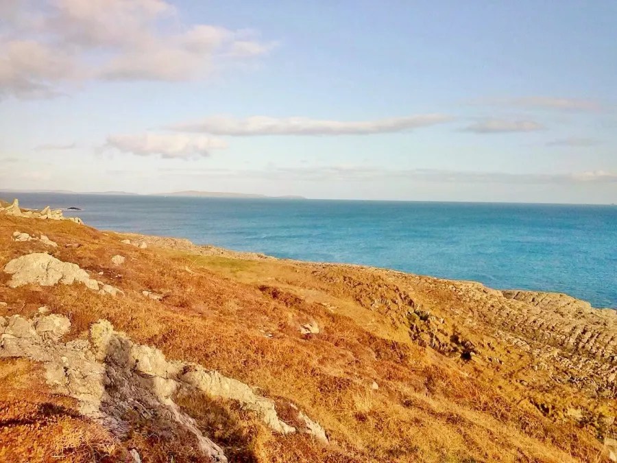 Weite Küstenlandschaft mit grünem Hügel und klarem blauem Wasser unter einem hellen Himmel.