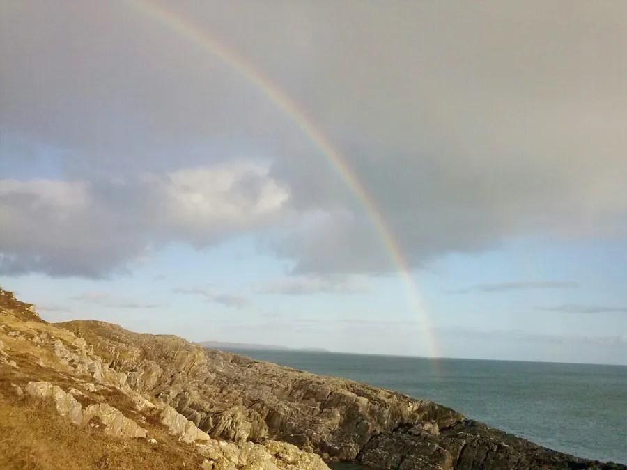 Ein Regenbogen spannt sich über eine felsige Küstenlandschaft mit Blick auf das Meer und eine helle Wolkenlandschaft.