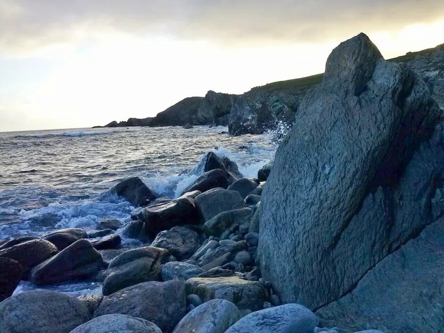 Eine felsige Küstenlinie mit Wellen, die gegen die Steine branden, unter einem bewölkten Himmel.