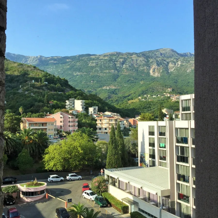 A view of a residential area in Tivat, Montenegro, featuring greenery, buildings, and mountains in the background under a clear blue sky.