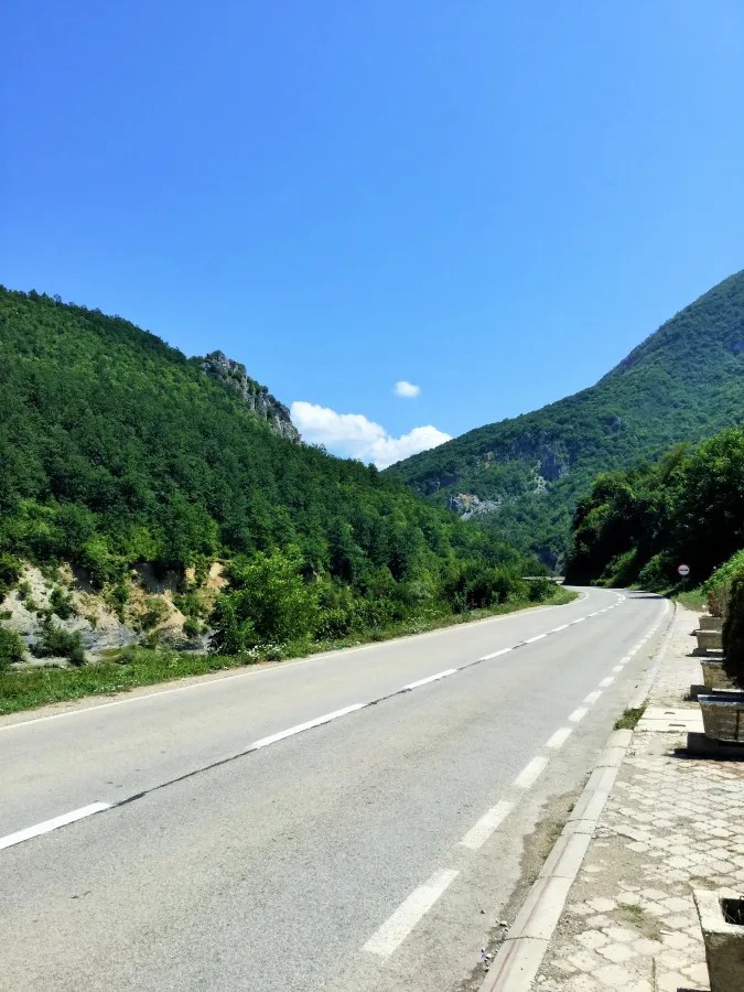 Landschaftsaufnahme einer kurvigen Straße umgeben von grünen Bergen und bewaldeten Hängen unter einem blauen Himmel.