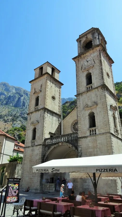 Blick auf das historische Gebäude mit zwei Türmen und einer Uhr im Herzen von Tivat, Montenegro. Im Vordergrund befindet sich ein Restaurant mit Tischen im Freien.