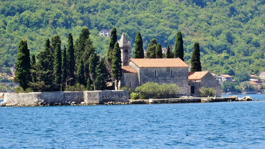 Ein Blick auf eine alte Kirche auf einer kleinen Insel, umgeben von hohen Zypressen und dem blauen Wasser einer Bucht, mit grünen Hügeln im Hintergrund.