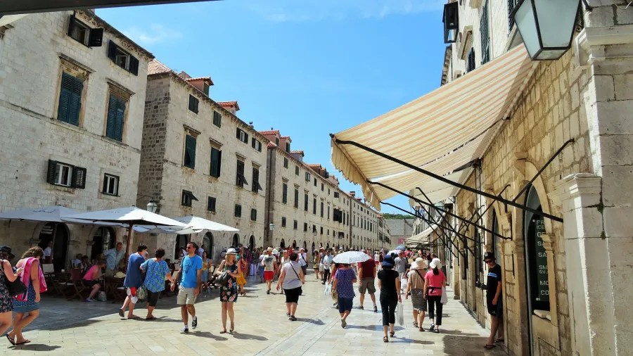 Belebte Straße in Dubrovnik mit historischen Gebäuden und Touristen, die sich unter Sonnenschirmen aufhalten.