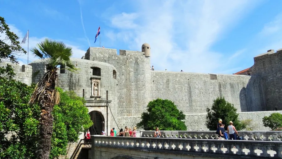 Blick auf die Stadtmauer von Dubrovnik mit Besuchern und Palmen im Vordergrund unter blauem Himmel.