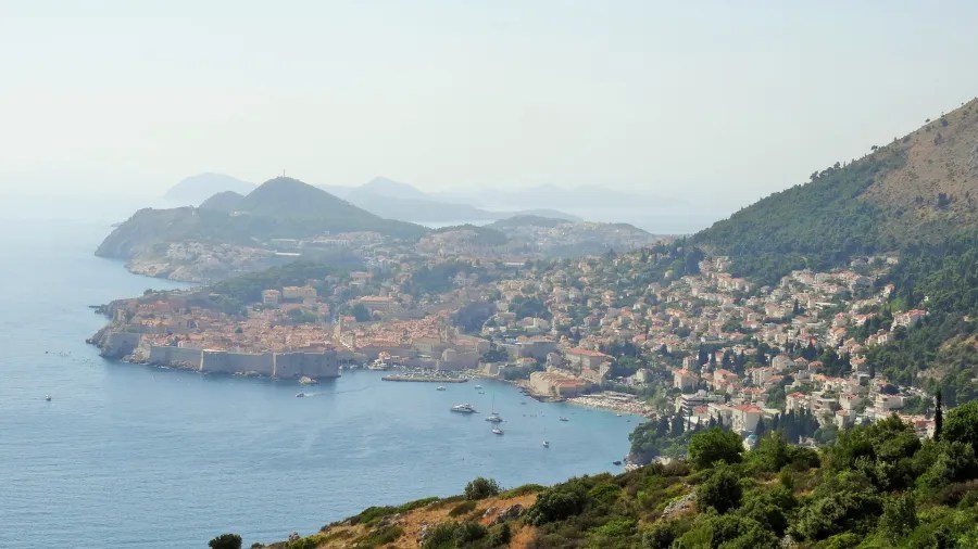 Panoramic view of Dubrovnik, Croatia, showcasing the historic city walls, houses, and the coastline, with mountains in the background.