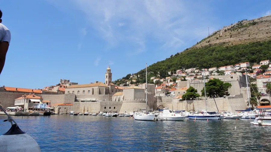 Blick auf die Altstadt von Dubrovnik mit historischen Gebäuden, Booten im Hafen und umliegenden Hügeln.