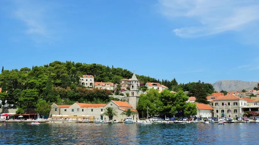 Panoramic view of a coastal village in Dubrovnik, Croatia, featuring orange-roofed buildings and lush greenery under a clear blue sky.