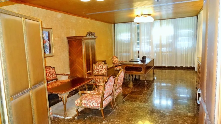 Innenansicht eines eleganten Raumes mit antiken Möbeln, darunter ein Schreibtisch und gepolsterte Stühle, umgeben von Fenstern mit weißen Vorhängen.