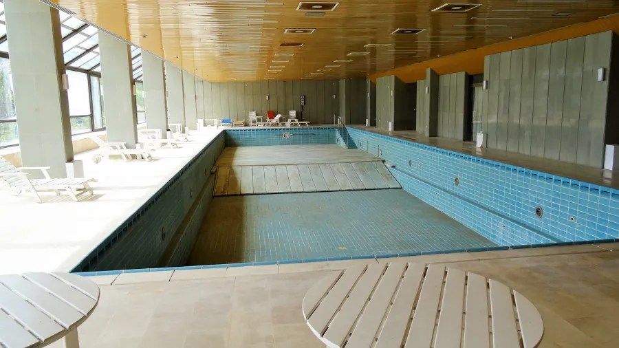 Innenansicht eines leeren Schwimmbads mit blauen Fliesen, Loungebereich und großen Fenstern.
