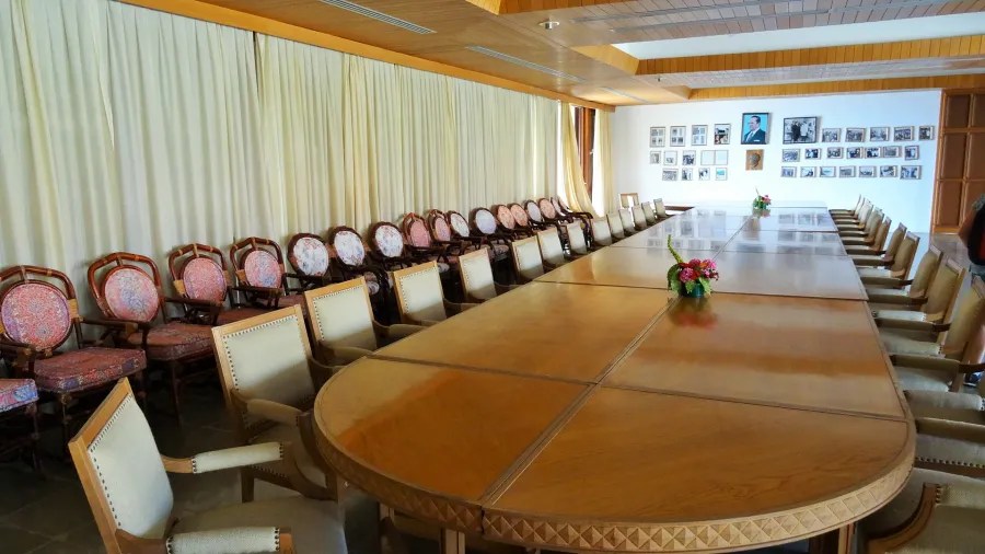 Innenansicht eines großen Konferenzraums mit einem langen Holztisch und Stühlen, umgeben von Vorhängen und historischen Fotografien an der Wand.