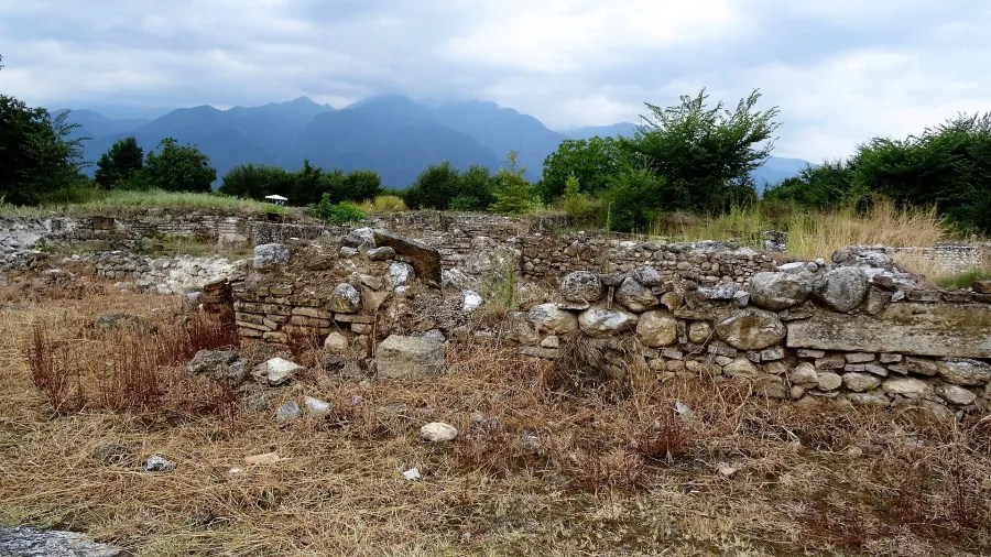 Ruinen aus Stein in einer trockenen Landschaft mit Hügeln im Hintergrund und bewachsenen Flächen.
