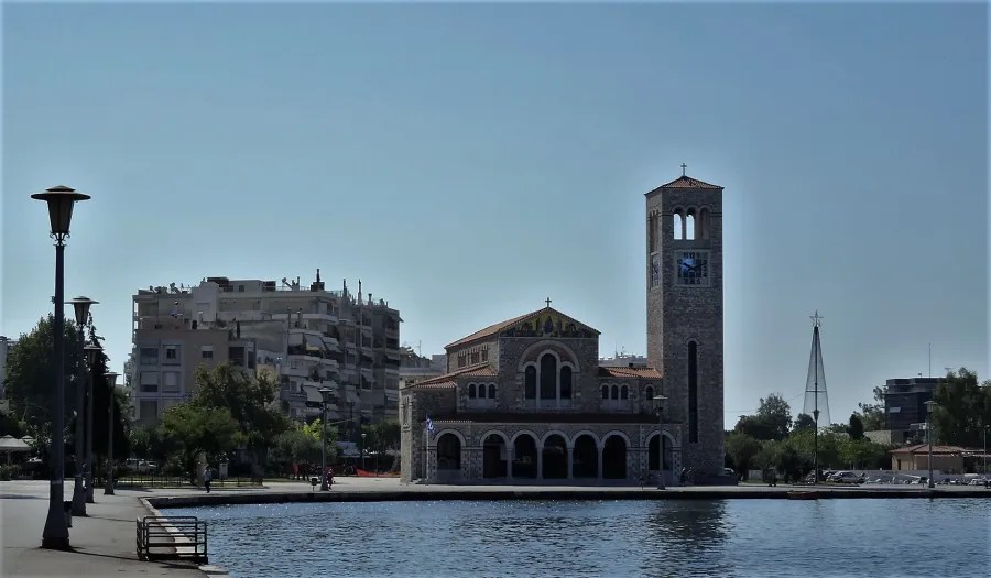 Blick auf eine Kirche mit einem hohen Glockenturm am Wasser in Griechenland, umgeben von städtischen Gebäuden und Bäumen, unter einem klaren blauen Himmel.