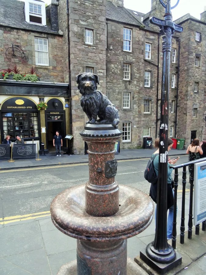 Bronzestatue eines Skye Terriers auf einem Denkmal, umgeben von historischen Gebäuden und einer belebten Straße in Edinburgh.