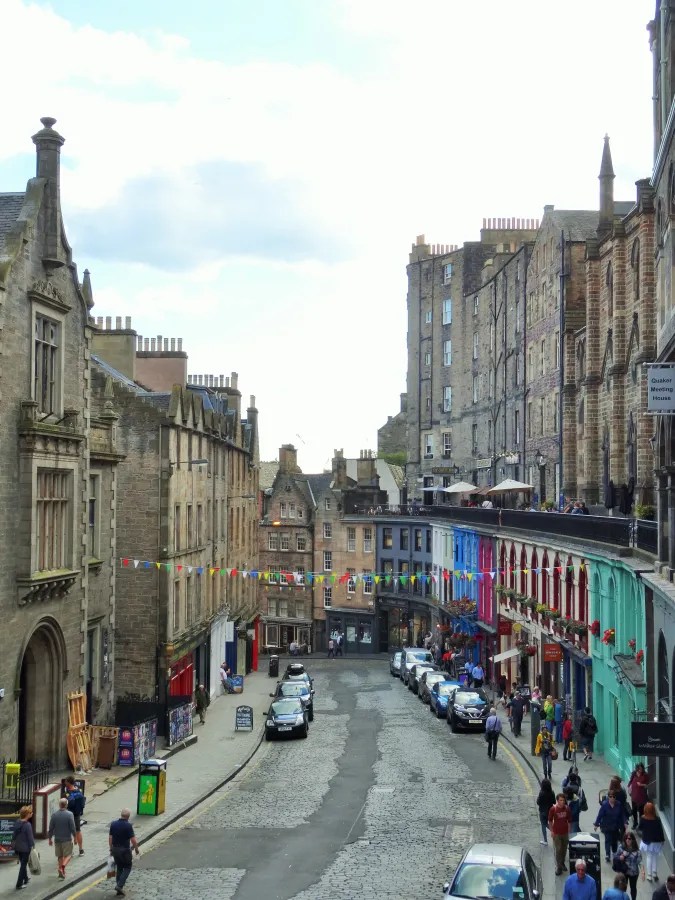 Eine belebte Straße in Edinburgh mit historischen Gebäuden und bunten Fassaden, gesäumt von Autos und Fußgängern, unter einem klaren Himmel.