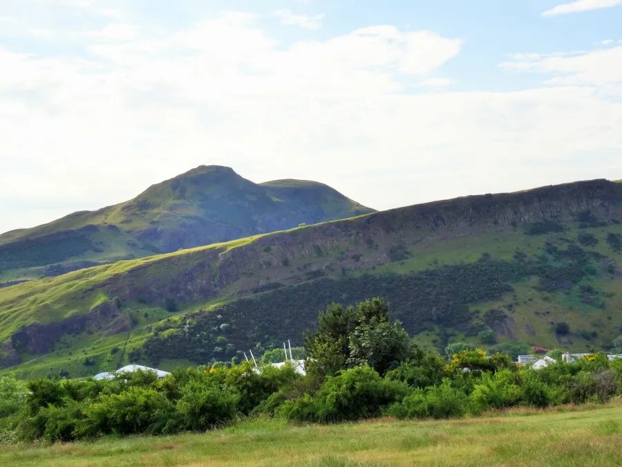 Blick auf die grünen Hügel und den Naturpark mit den Salisbury Crags und Arthur's Seat in Edinburgh unter einem blauen Himmel.