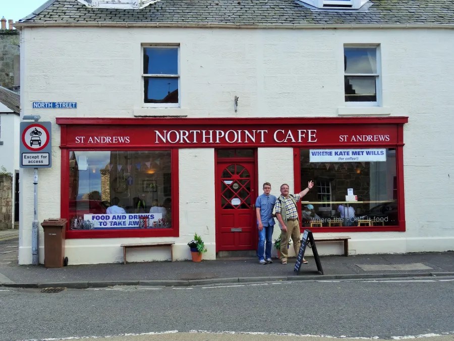 Northpoint Café in St. Andrews, featuring a red exterior with a sign indicating its historical connection to Kate and William. Two men stand in front, smiling and posing for the photo.