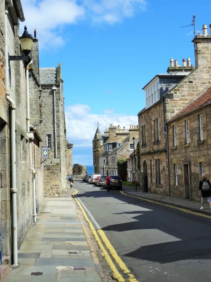 Blick auf eine charmante Straße in St. Andrews, Schottland, mit alten Steinbauten und parkenden Autos. Der Ozean ist in der Ferne sichtbar unter einem strahlend blauen Himmel.