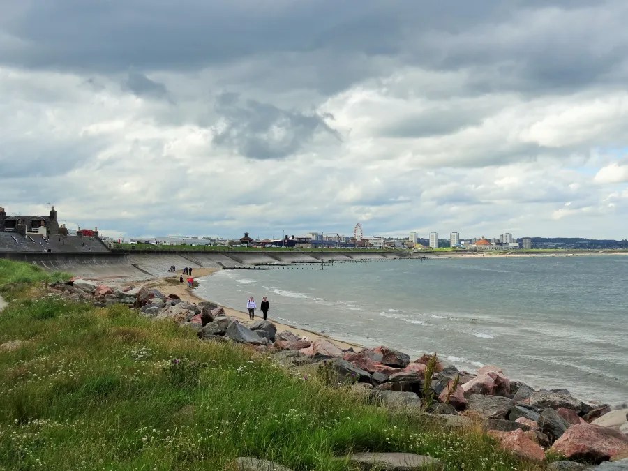 Blick auf die Küste mit Gras, Steinen und Menschen, die am Strand spazieren, während Wolken am Himmel hängen.