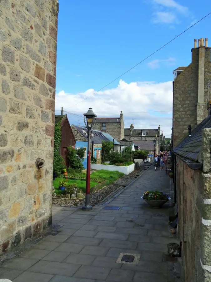 Ein malerischer Gang in Footdee, einem kleinen Stadtteil von Aberdeen, mit historischen Steinhäusern und einem blauen Himmel im Hintergrund.