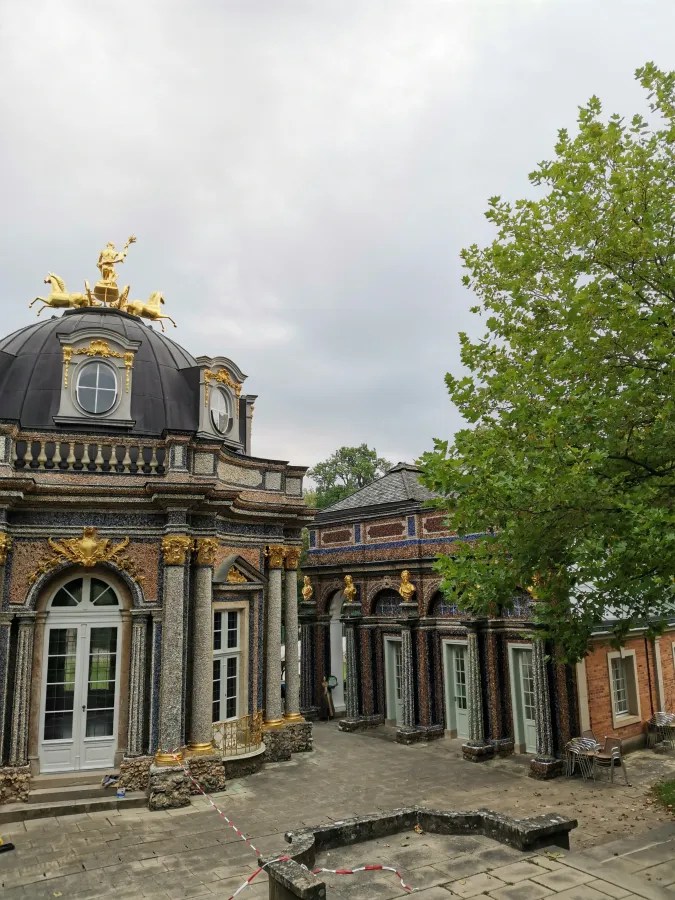 Ansicht des barocken Gebäudes mit goldenen Verzierungen und buntem Mosaik, umgeben von Bäumen und unter einem grauen Himmel.