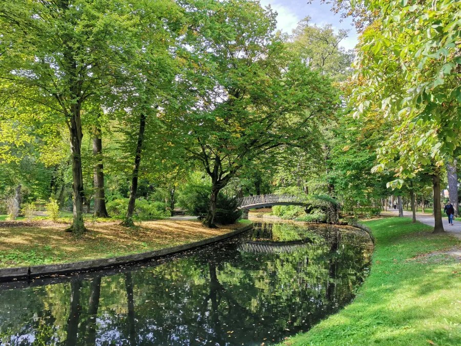 Ein malerischer Park mit grünem Laub, Bäumen und einem ruhigen Kanal, an dem eine kleine Brücke zu sehen ist.